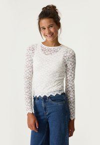 Weißes langärmliges Oberteil aus Spitze mit Blumenmustern, Rundhalsausschnitt und gewelltem Saum, kombiniert mit blauen Denim-Jeans.