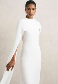 Robe blanche longue jusqu'au sol avec un col haut et des manches en cape fluides, présentant une texture lisse et une silhouette ajustée. Pas de motifs visibles.