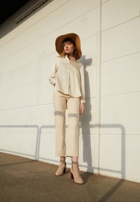 Chemise en soie crème à boutons, pantalon évasé beige, bottines marron à talon bloc, et un chapeau de paille à large bord contre un mur blanc.
