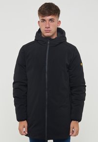 Parka nera imbottita con cappuccio, zip frontale eTaspringo. Dotata di un distintivo logo giallo sulla manica sinistra. Texture liscia.