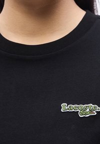 Gros plan d'une personne portant une chemise noire avec un logo Lacoste vert représentant un crocodile sur le côté gauche de la poitrine.