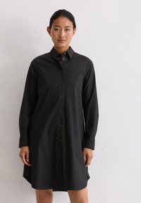 Femme posant contre un mur blanc, portant une robe chemise noire à manches longues avec une poche poitrine et un col.