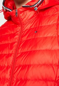 Červená péřová bunda se zipem vpředu, vysokým límcem a nastavitelnou kapucí. Obsahuje malé logo na levé straně hrudi a horizontální prošití.