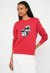 Nő piros pulóvert visel, amelyen egy fekete-fehér rajzfilmfigura látható, rajta napszemüveggel, valamint egy macska, amely szintén napszemüveget visel, fehér szoknyában, fehér háttér előtt áll.