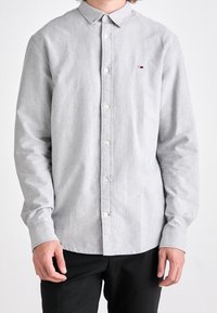 Uomo che indossa una camicia a maniche lunghe grigia chiaro con bottoni e pantaloni neri, in piedi con le braccia rilassate ai lati su uno sfondo semplice.