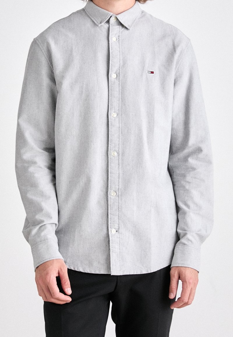 Uomo che indossa una camicia a maniche lunghe grigia chiaro con bottoni e pantaloni neri, in piedi con le braccia rilassate ai lati su uno sfondo semplice.