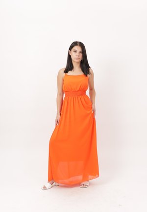Joven mujer con pelo liso y oscuro que lleva un vestido largo sin mangas de color naranja brillante y tacones blancos de punta abierta, de pie sobre un fondo blanco.