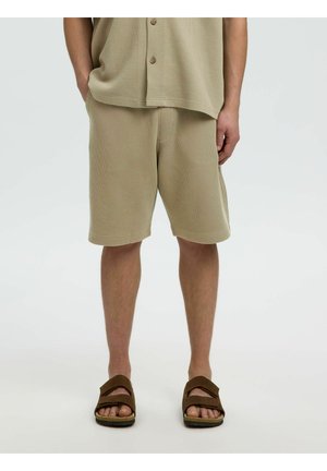 Mężczyzna ubrany w beżowe, teksturowane szorty i pasującą koszulę z krótkim rękawem, stoi z rękami częściowo w kieszeniach, ma na nogach brązowe sandały.