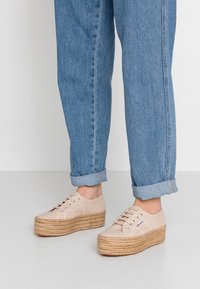 Sneakers platform in tela beige con suole in stile espadrilla a intreccio. Presentano cinque asole e lacci regolabili. Indossati con jeans blu arrotolati.