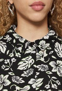 Svart blommig blus med krage som har vita och gröna bladmönster, knäppning fram och ett mjukt, texturerat tyg.