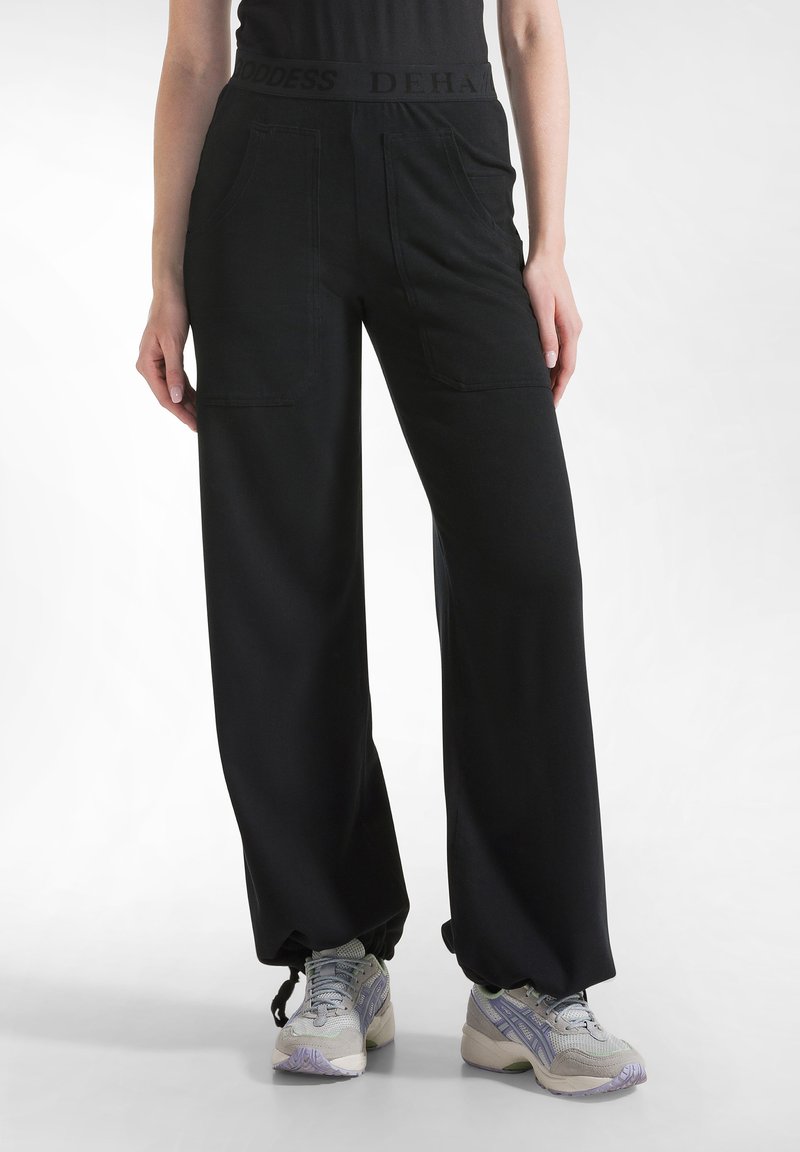 Zwarte wijde broek met een hoge tailleband met de tekst "GODDESS DEHA." De broek heeft voorzakken en een gladde, zachte textuur.