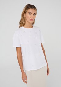 T-shirt bianco a maniche corte con scollatura rotonda, impreziosito da dettagli floreali e perline sulla parte anteriore. Realizzato in morbido cotone.