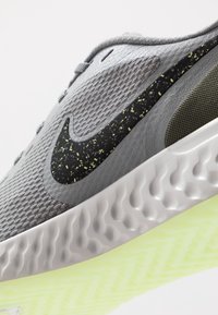 Baskets de course Nike avec une tige en maille grise, des accents noirs légèrement pigmentés de jaune, et une semelle blanche texturée avec une base verte translucide.