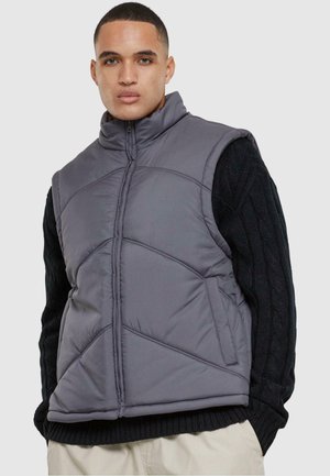 Grå puffer vest med høj krave og lynlås, med quiltet design og to sidelommer. Båret over en tekstureret sort sweater.