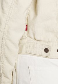 Vit kordjacka med synliga sömmar, med en röd Levi's etikett och bronsknappar. Inkluderar en framficka och textil med struktur.