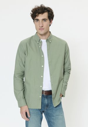 Lys grøn bomuldsskjorte med knapper, båret over en hvid t-shirt. Lange ærmer, enkelt brystlomme, afslappet pasform, kombineret med blå jeans.