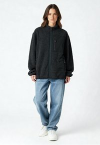 Veste en polaire noire avec fermeture éclair devant, comprenant une poche à panneau noir uni, portée sur un jean bleu clair et des baskets blanches.