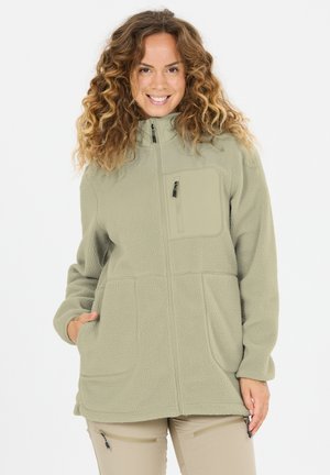 Frau mit lockigem Haar, die lächelt, trägt eine beige Fleecejacke mit Reißverschlusstaschen an Brust und Seiten, steht vor einem schlichten weißen Hintergrund.