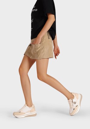 Mini gonna in suede beige con tasche laterali, abbinata a sneakers crema e beige con inserti in rete, dettagli metallici e suola piatta.
