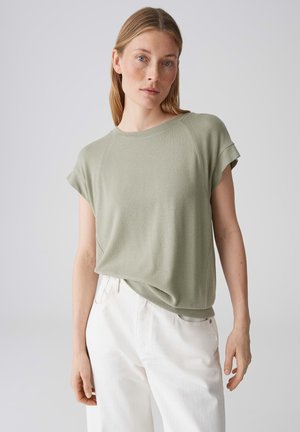 Donna con capelli biondi lisci indossa una maglietta verde chiaro a maniche corte e pantaloni bianchi a vita alta, in piedi davanti a uno sfondo grigio uniforme.