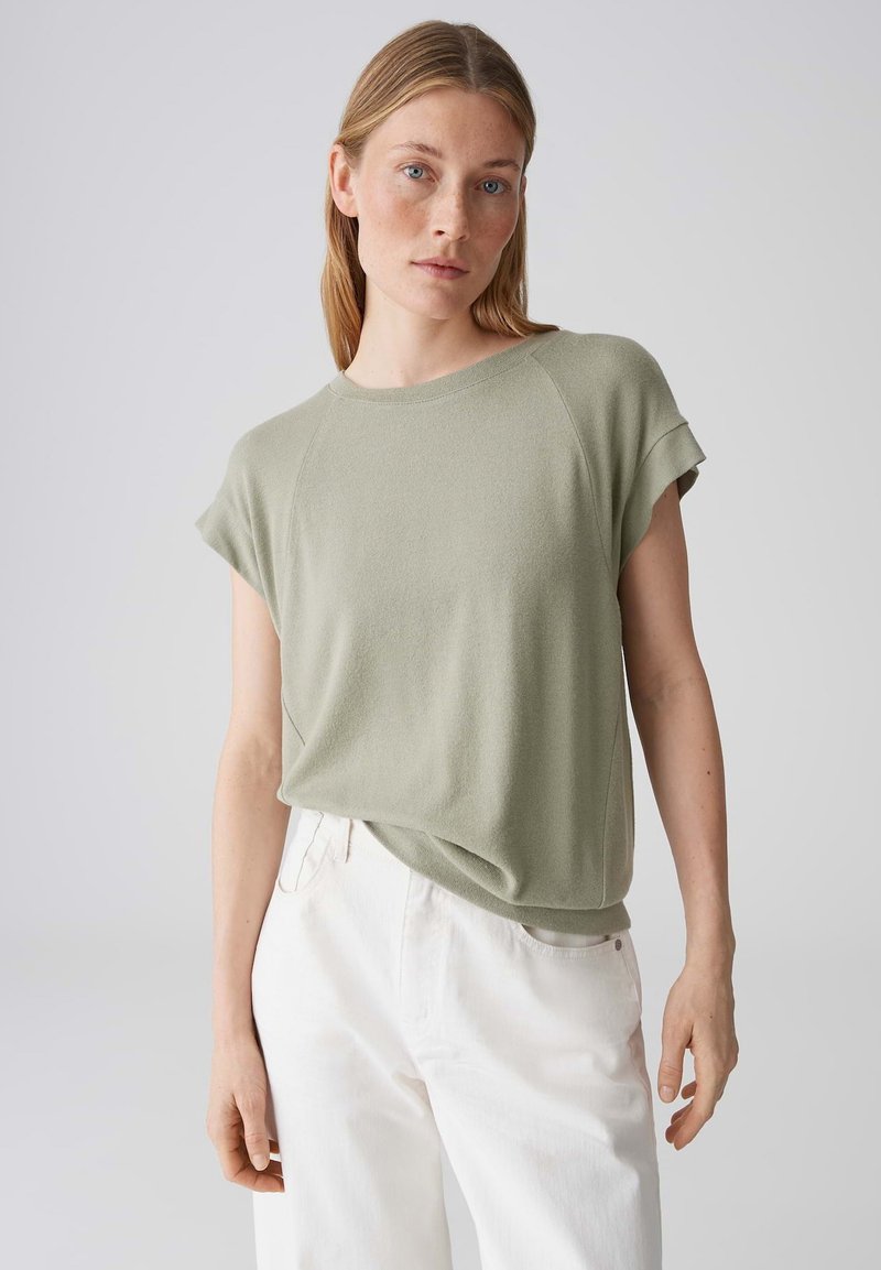 Femme aux cheveux blonds lisses portant un haut vert clair à manches courtes et un pantalon blanc taille haute, debout devant un fond gris uni.