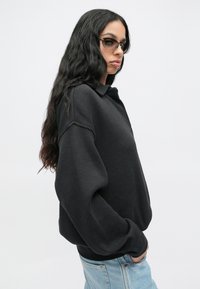 Mulher com cabelo longo e ondulado, usando óculos de sol escuros, um suéter oversized preto texturizado e jeans azul claro, posando com as mãos nos bolsos.