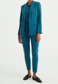 Modrý blazer s klopovým límcem, jednobuttonovým zapínáním, odpovídajícími tapered kalhotami a přiléhavým tmavě modrým svetrem. Černé mokasíny dotvářejí tento outfit.