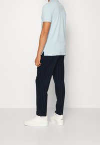 Man som bär en ljusblå kortärmad polo, marinblå randiga byxor, vita strumpor och vita sneakers, stående mot en enfärgad vit bakgrund.