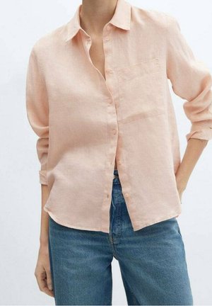 Femme portant une chemise pêche clair à manches retroussées et un jean bleu taille haute, debout avec une main dans la poche.