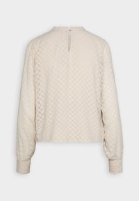 Blouse beige clair avec un motif en losanges texturé. Elle présente de longues manches bouffantes et un décolleté au dos avec une fermeture à bouton unique.