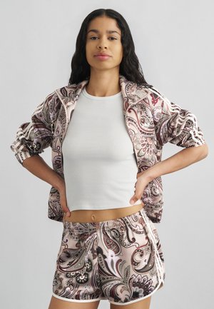 Mujer con cabello largo y oscuro, vestida con un top corto blanco, chaqueta con estampado de cachemira y pantalones cortos a juego, de pie con las manos en las caderas frente a un fondo gris.