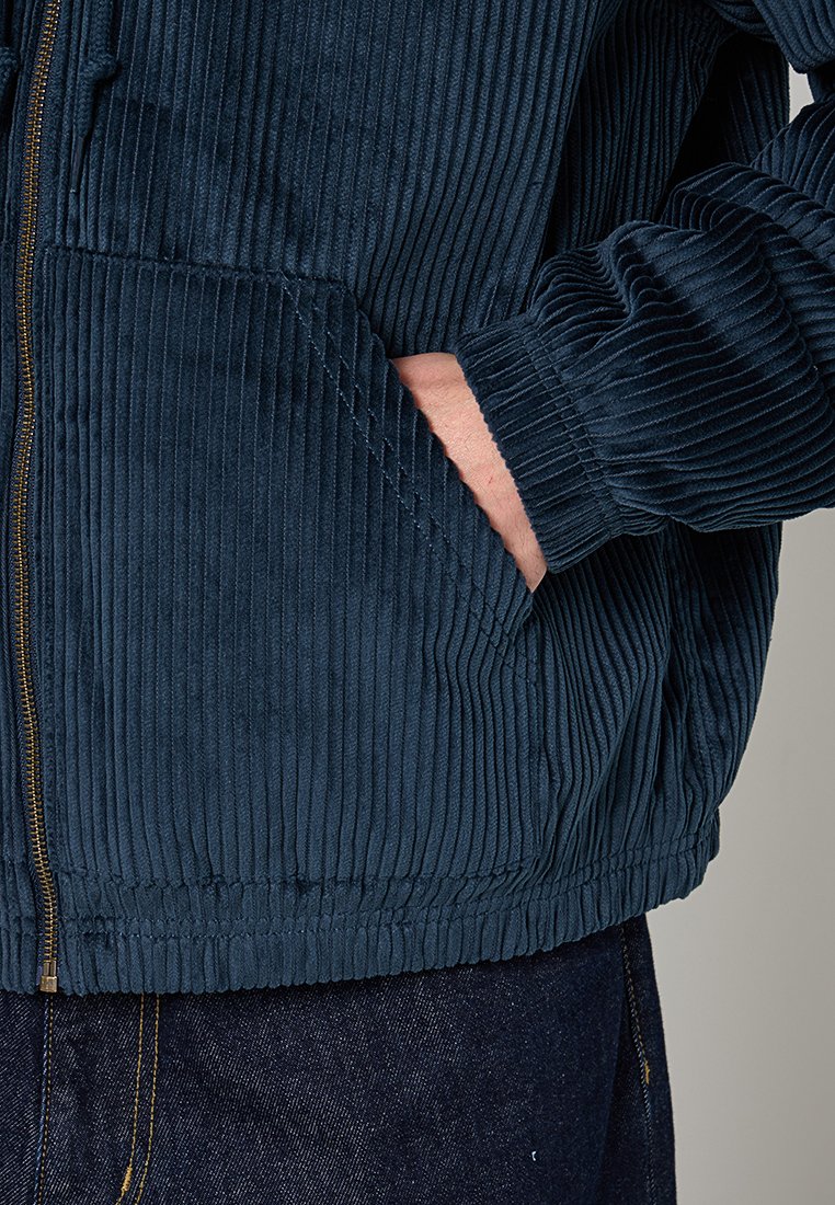 Veste en velours côtelé bleu marine avec des côtes verticales, fermeture éclair et poches latérales. Les poignets sont côtelés ; le tissu a une finition douce et texturée.