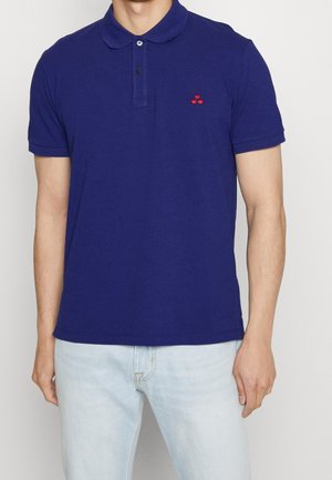 Mężczyzna ubrany w dopasowaną granatową koszulkę typu polo z krótkim rękawem i jasnoniebieskie dżinsy, stojący z ramionami swobodnie opuszczonymi wzdłuż ciała.