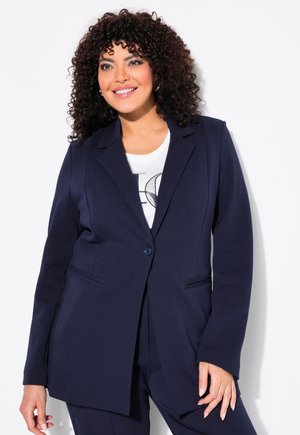 Blazer lungo blu navy in tessuto liscio con chiusura a un bottone, revers con punte e tasche laterali, abbinato a una t-shirt bianca con grafica.