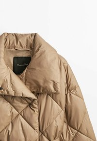 Veste matelassée beige avec col large et rembourré, fermeture par boutons pression, présentant une texture de tissu lisse et brillante.