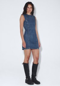 Denimmekko, jossa on hihaton muotoilu, sininen väri, näkyvät ompelutiedot ja suora leikkaus. Yhdistetty polven yli ulottuviin mustiin nahkabootseihin.