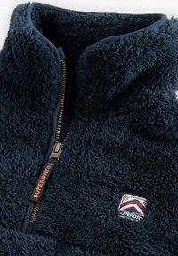 Veste en polaire bleu marine avec un col haut, fermeture éclair dorée et tissu texturé ressemblant à de la fourrure. Présente une pièce de logo rouge et blanc sur la poitrine.