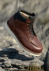 Brauner Lederstiefel mit abgerundeter Zehenpartie, schwarzem gepolstertem Kragen, weißer Gummisohle und gestickten Details. Schwebt über felsigem Terrain.