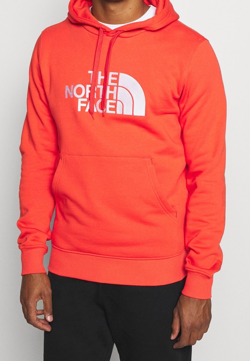 Personne portant un sweat à capuche orange vif avec le logo blanc "The North Face", pantalon noir, debout devant un fond clair uni.