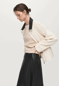 Giacca crema con colletto nero, maniche lunghe e zip. Indossata sopra una camicia beige e una gonna di pelle nera. Texture lisce.