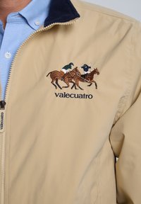 Chaqueta beige con un cuello azul marino, que presenta una escena bordada de tres jinetes. El nombre de la marca "valecuatro" se muestra debajo.