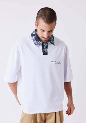 Homme aux cheveux courts portant une chemise blanche à manches courtes avec un col sombre à motif et un pantalon beige, regardant vers le bas.