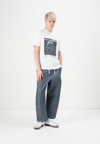 Young man with short blonde hair wears white graphic tee, loose blue pants, and white sneakers, standing against a plain white background.