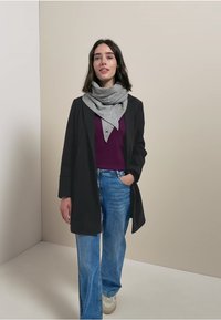 Cappotto lungo nero sopra una maglietta viola, abbinato a jeans azzurri e una sciarpa grigia. Il modello è in piedi contro uno sfondo neutro.