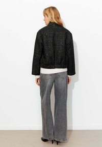 Veste courte texturée noire avec un col montant, une taille élastique et des manches longues, portée par-dessus un pull blanc et un jean gris à jambes larges.