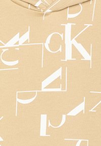 Felpa beige con grandi lettere bianche in stile blocco disposte in varie orientazioni e dimensioni su tutta la stoffa. Materiale in cotone con una consistenza morbida.