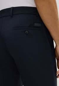 Marineblaue Hose mit glatter Oberfl�äche, ausgestattet mit einer Gesäßtasche mit Knopf und einem kleinen Markenlabel. Detaillierte Nähte betonen die Säume.