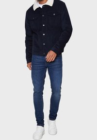 Giacca in velluto a coste navy con colletto in pelliccia artificiale bianca, bottoni a scatto sul davanti e due tasche sul petto. Abbinata a jeans slim-fit blu e sneakers bianche.