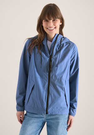 Młoda kobieta uśmiechająca się, ubrana w niebieską przeciwdeszczową kurtkę z kapturem i jasnoniebieskie dżinsy na jednolitym tle.