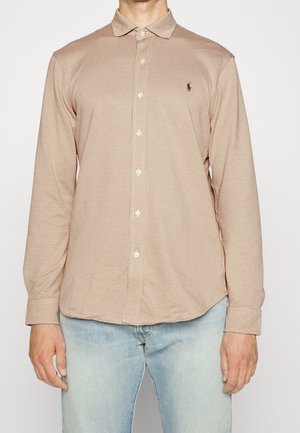 Mand iført beige ternet skjorte med lange ærmer og krave og lyseblå jeans, stående mod en ensfarvet baggrund.